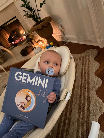 Baby holding a book titled 'Gemini Baby' in a living room setting.
