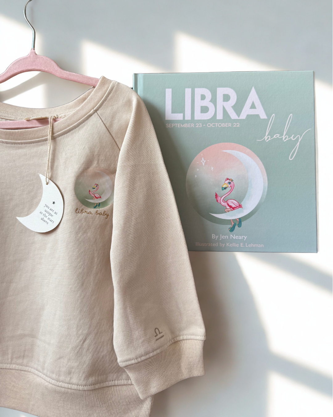 A beige crewneck sweatshirt with a flamingo graphic on the chest hangs on a pink hanger next to a printed book or card titled 'LIBRA baby' featuring a flamingo illustration, set against a white wall with soft sunlight shadows.